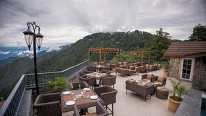 An outdoor patio with wicker furniture, tables set for dining, and a pergola with string lights. -  Presidium Sarovar Premiere Dalhousie (Dalhousie)