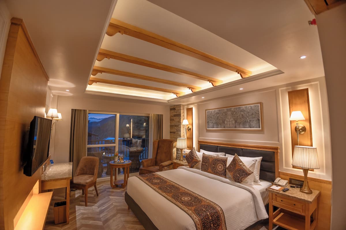 A hotel room with a large bed, a TV, a desk, and a chair. There is a window with a view of the mountains.