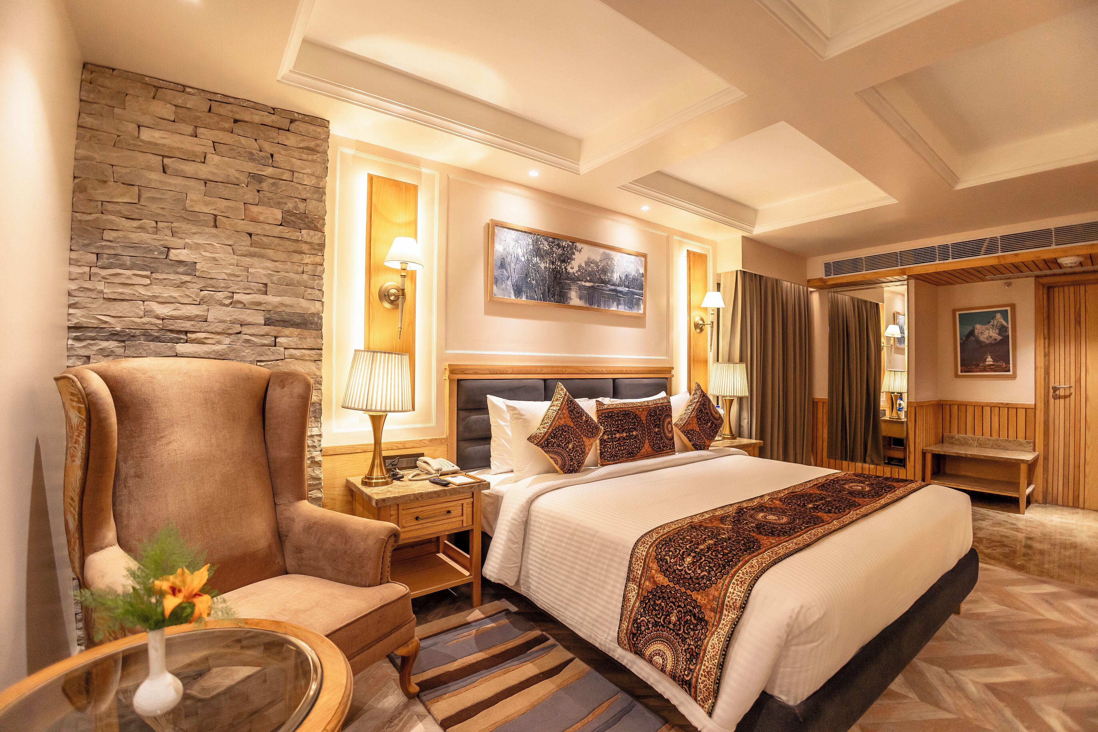 Une chambre d’hôtel avec un grand lit, un mur décoratif en pierre, un banc en bois et une table de chevet avec une lampe.