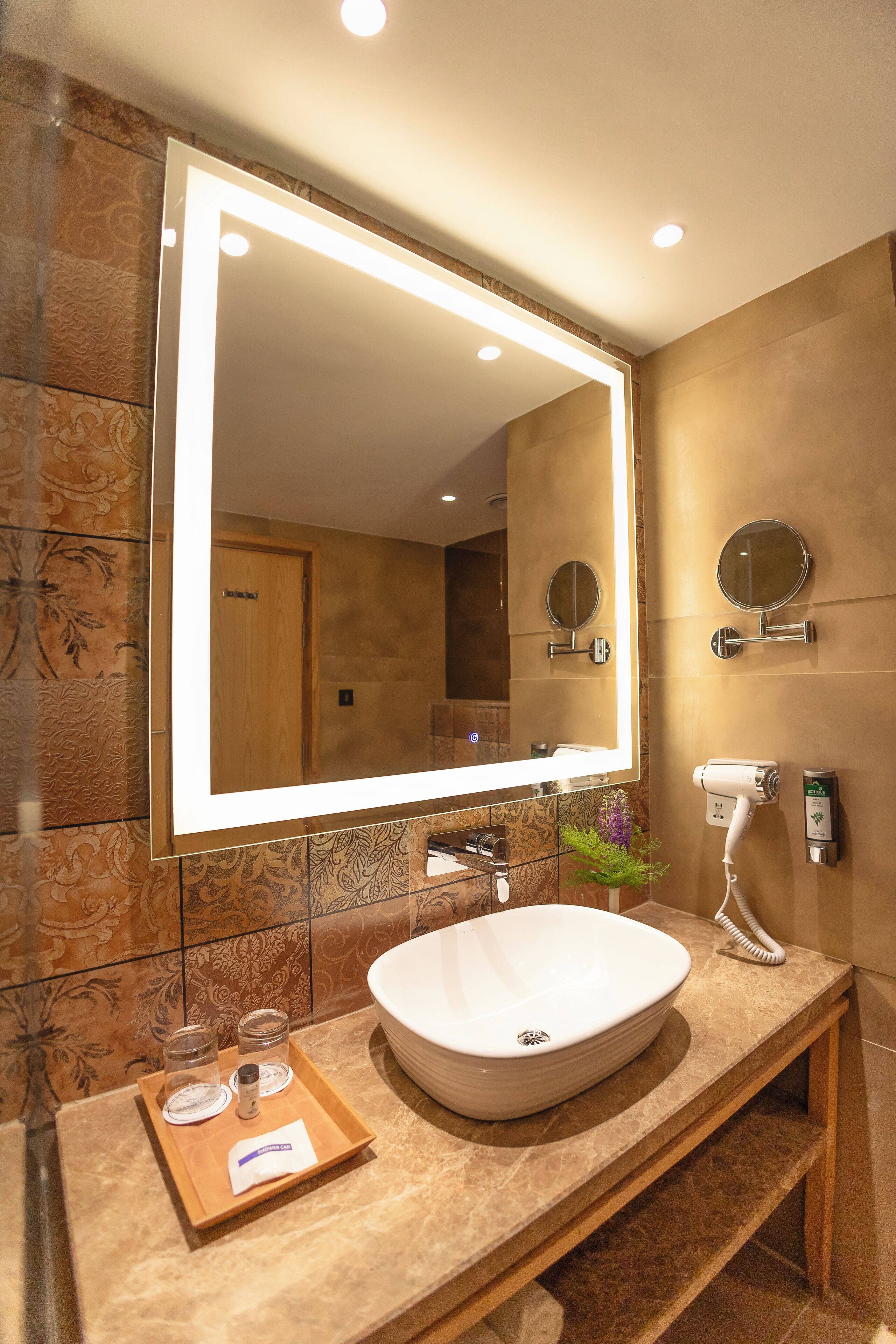 A bathroom with a large mirror, a white sink, a hairdryer, and a tray with toiletries.