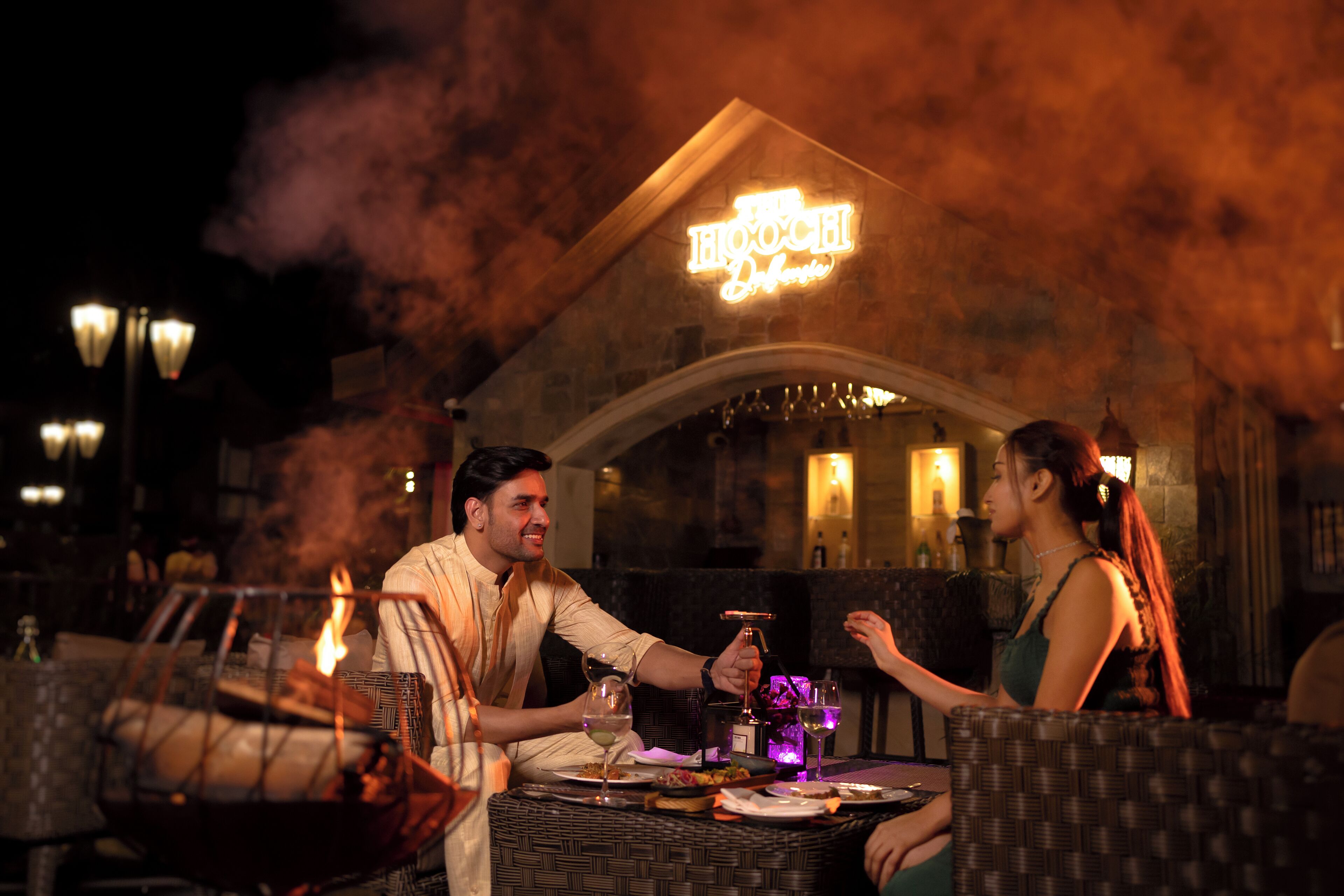 Un restaurante con un letrero de neón, mesas al aire libre y una chimenea.