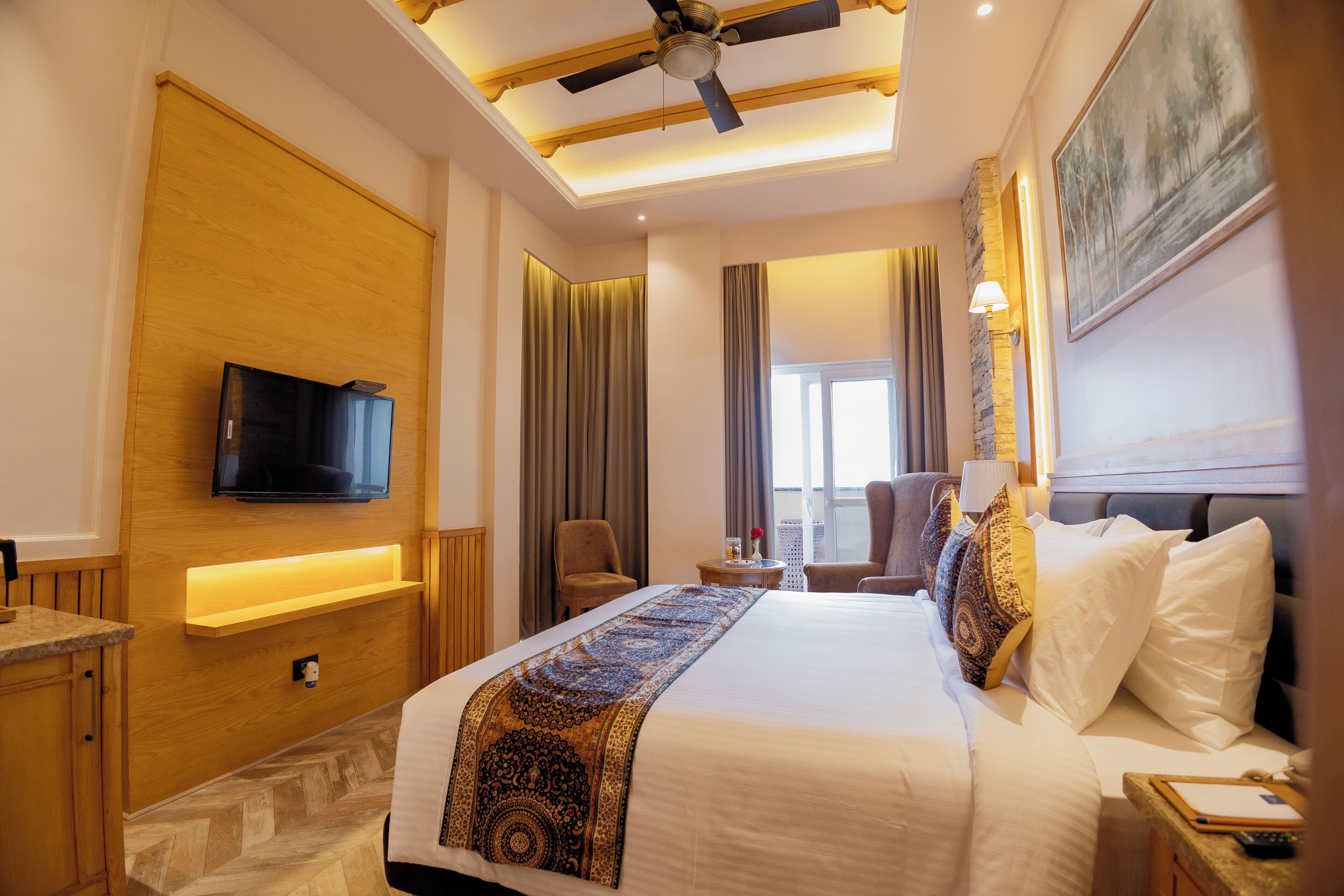 A hotel room with a large bed, a television mounted on the wall, a ceiling fan, and a window with curtains.