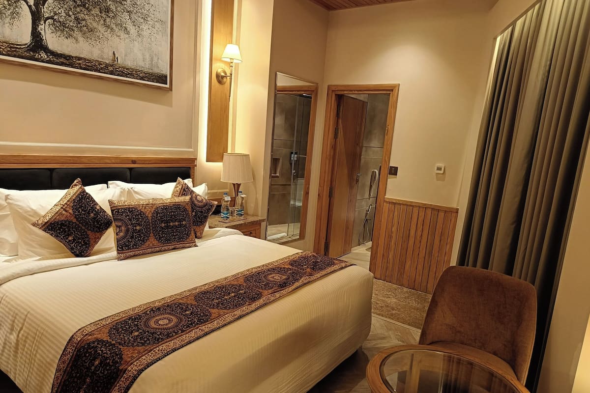 A hotel room with a large bed, a chair, a glass table, a bathroom with a shower, and a painting on the wall.