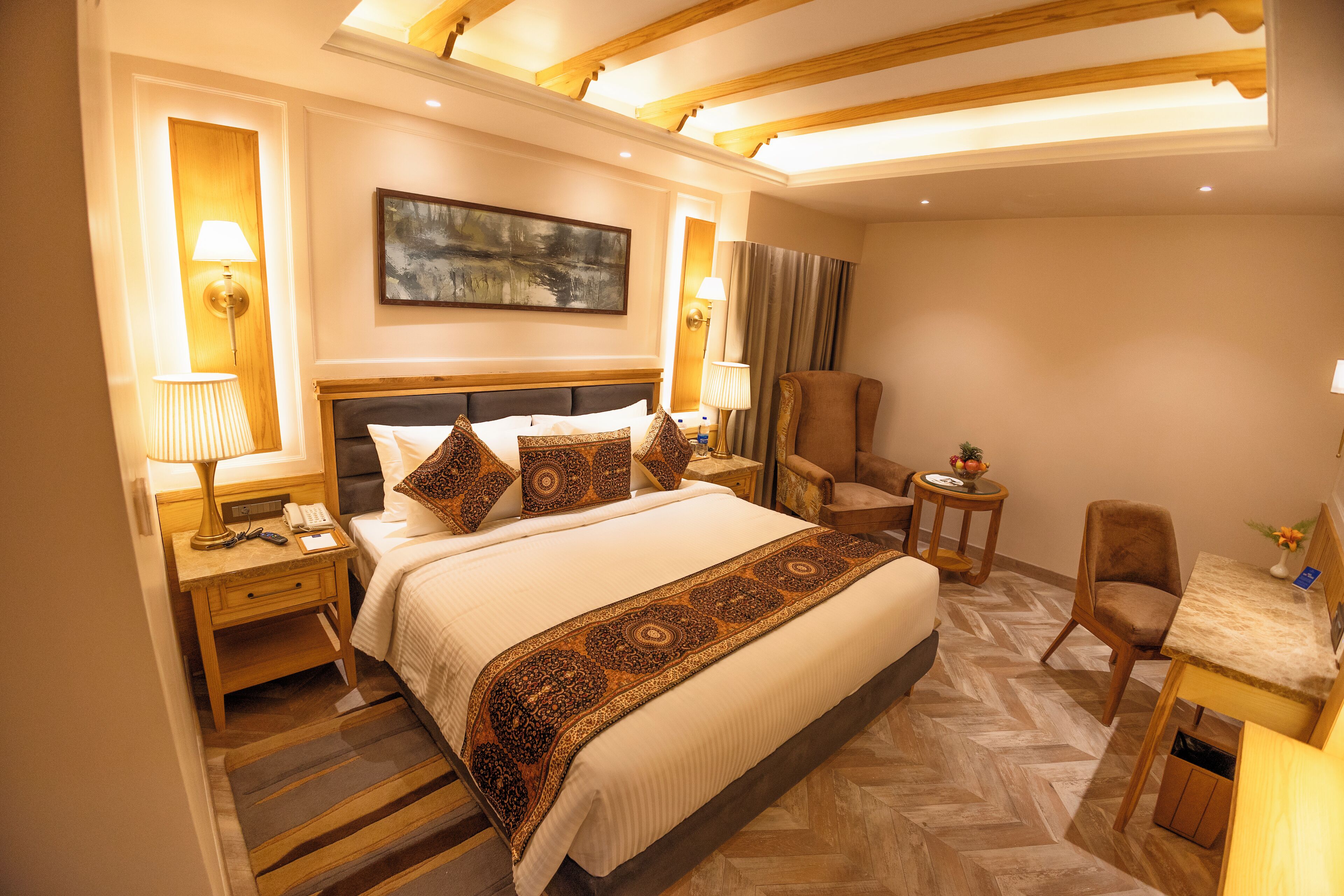 Une chambre d’hôtel avec un grand lit, deux tables de chevet avec des lampes, un bureau avec une chaise, un tableau au mur et un parquet à motifs en chevrons.