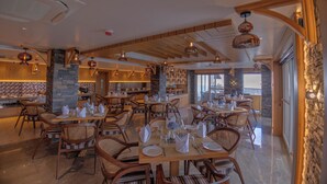 A spacious restaurant with wooden tables and chairs, stone pillars, and hanging pendant lights. -  Presidium Sarovar Premiere Dalhousie (Dalhousie)