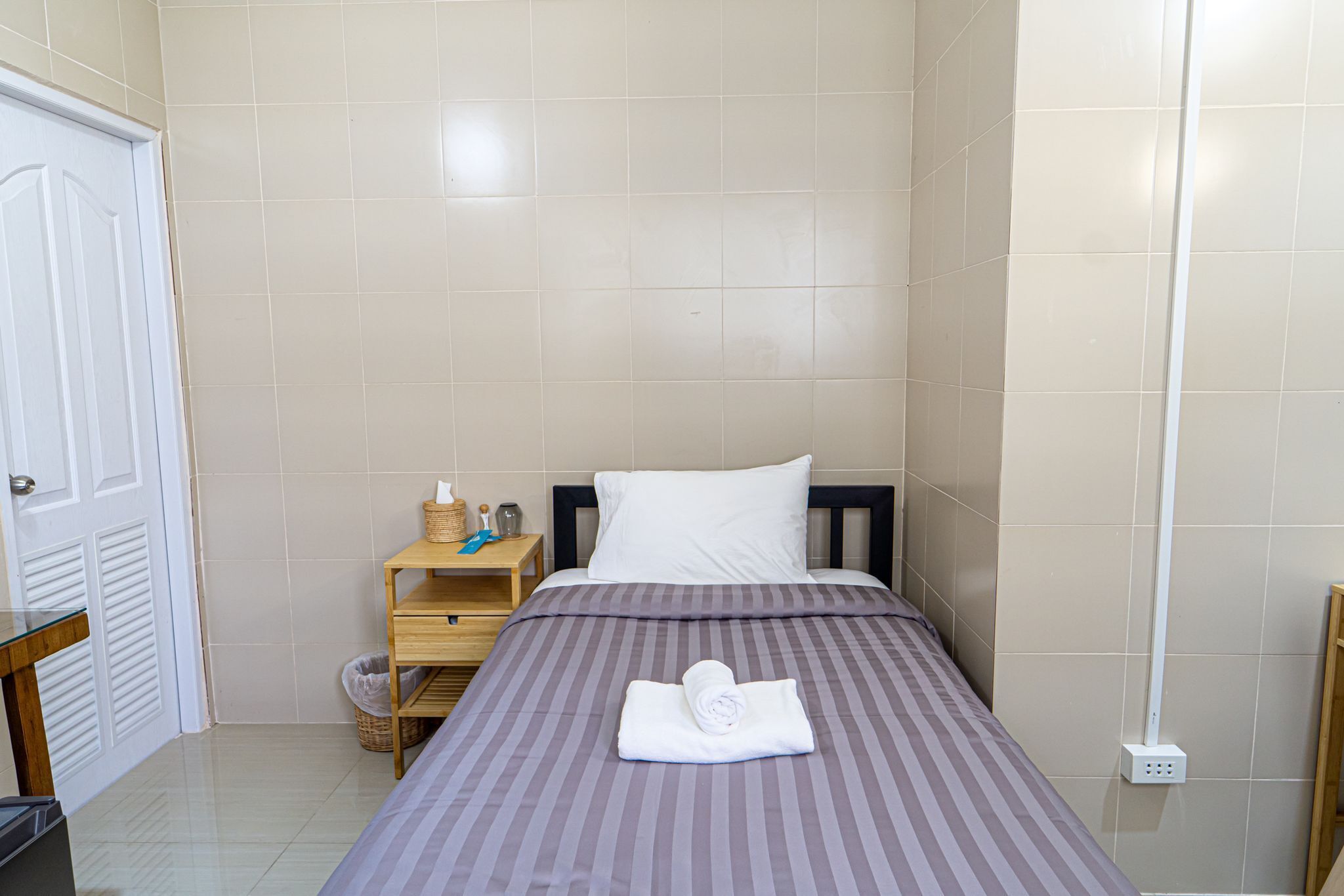 A neatly made bed with a striped bedspread and a rolled towel on top. There is a wooden nightstand with a lamp, a basket, and a small table beside the bed.