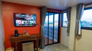 A hotel room with a large TV displaying a streaming service, a wooden desk, a mini-fridge, a wicker basket, and a balcony with a view of traditional buildings. - Lo-ha Guesthouse Contactless Check-in (Bangkok)