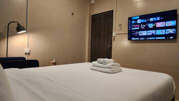 A hotel room with a bed, a TV mounted on the wall displaying various channels, and a dark wooden door.
