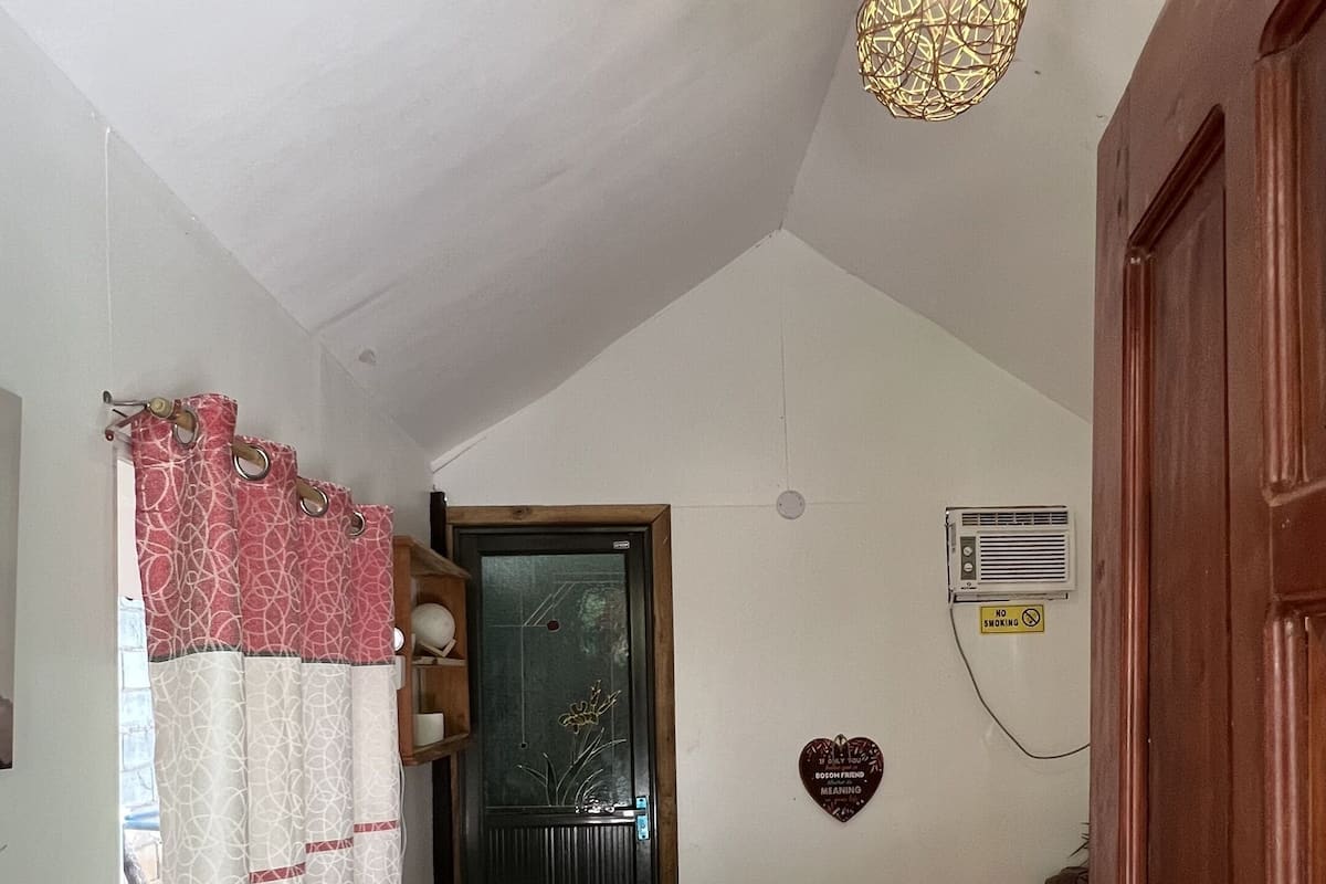 Um quarto com porta de madeira, uma cama, janela com cortinas, aquecedor fixado na parede e luminária no teto.