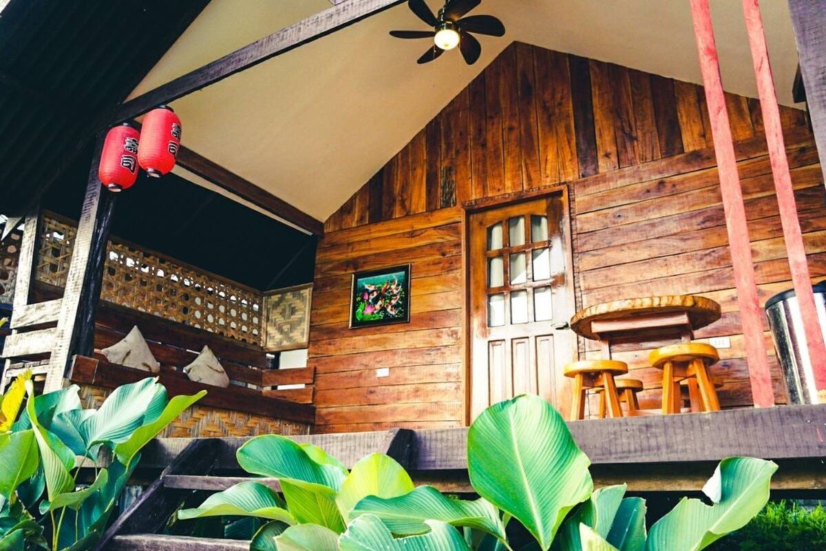Uma cabana de madeira com ventilador de teto, porta de madeira e uma pequena mesa com banquinhos.