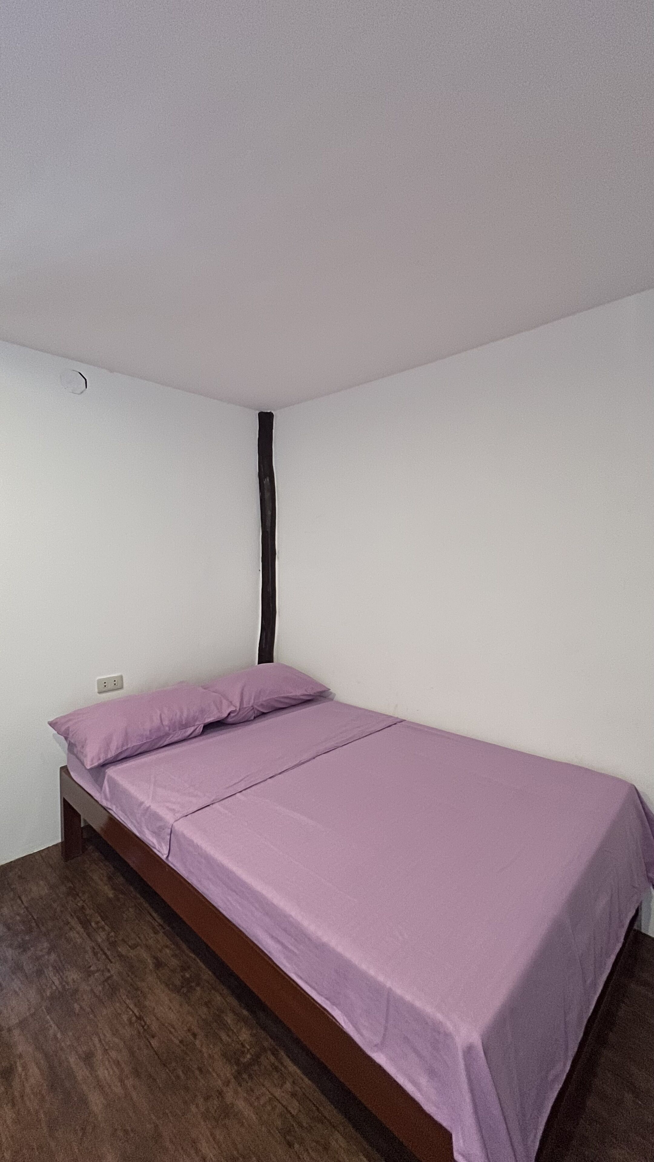 A small, single-bed room with a wooden frame, a purple bedspread, and a white wall.