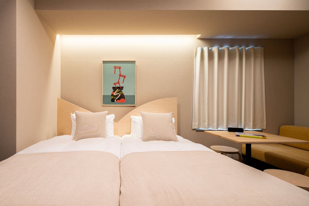 A hotel room with two beds, a desk, a chair, a painting on the wall, and a window with curtains.