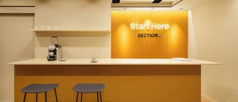 A reception area with a counter, two bar stools, a coffee machine, and a sign that reads 'Start Here SECTION L'.