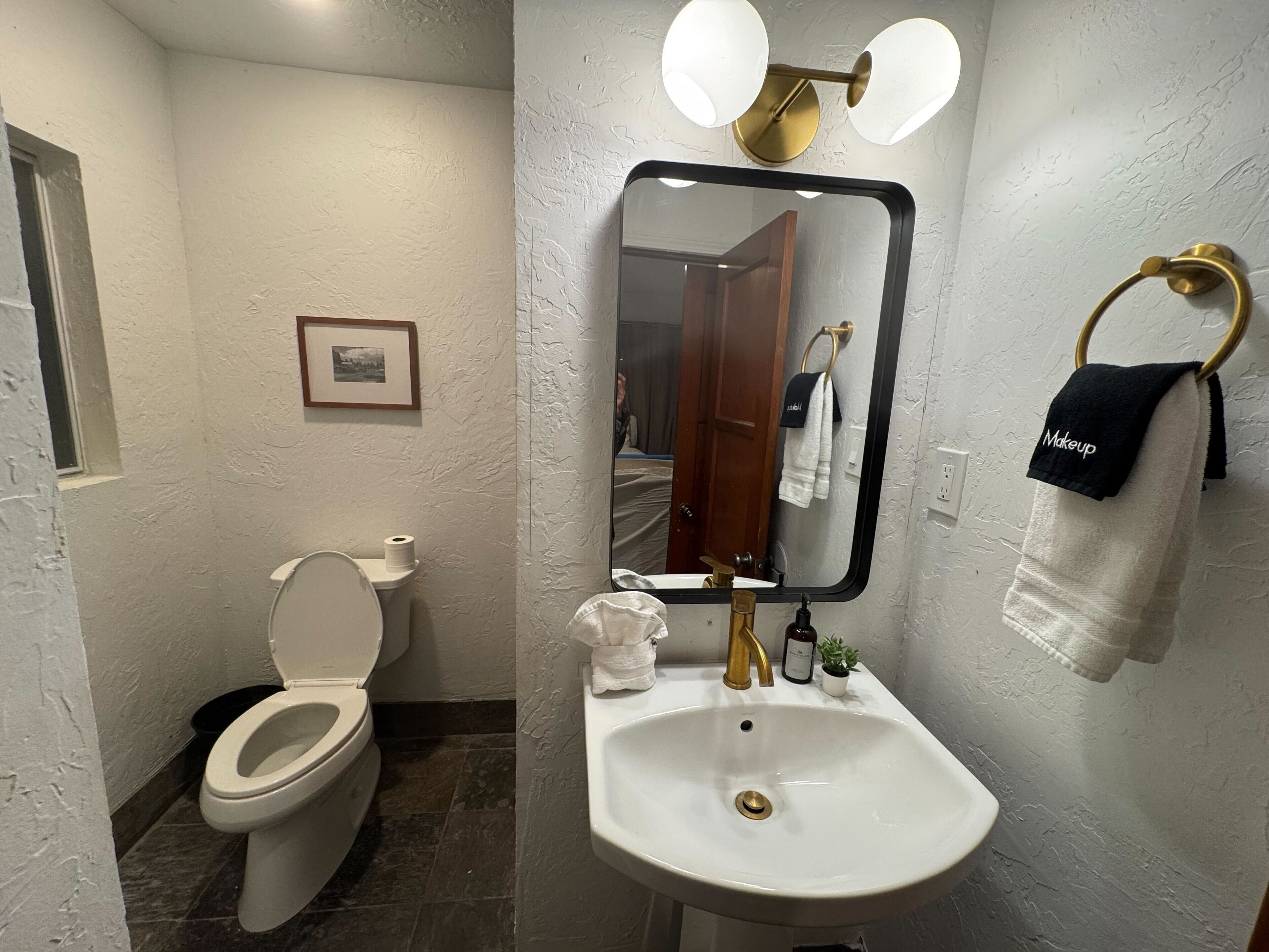 A bathroom with a toilet, sink, and mirror. There is a towel rack with towels and a small potted plant on the sink.