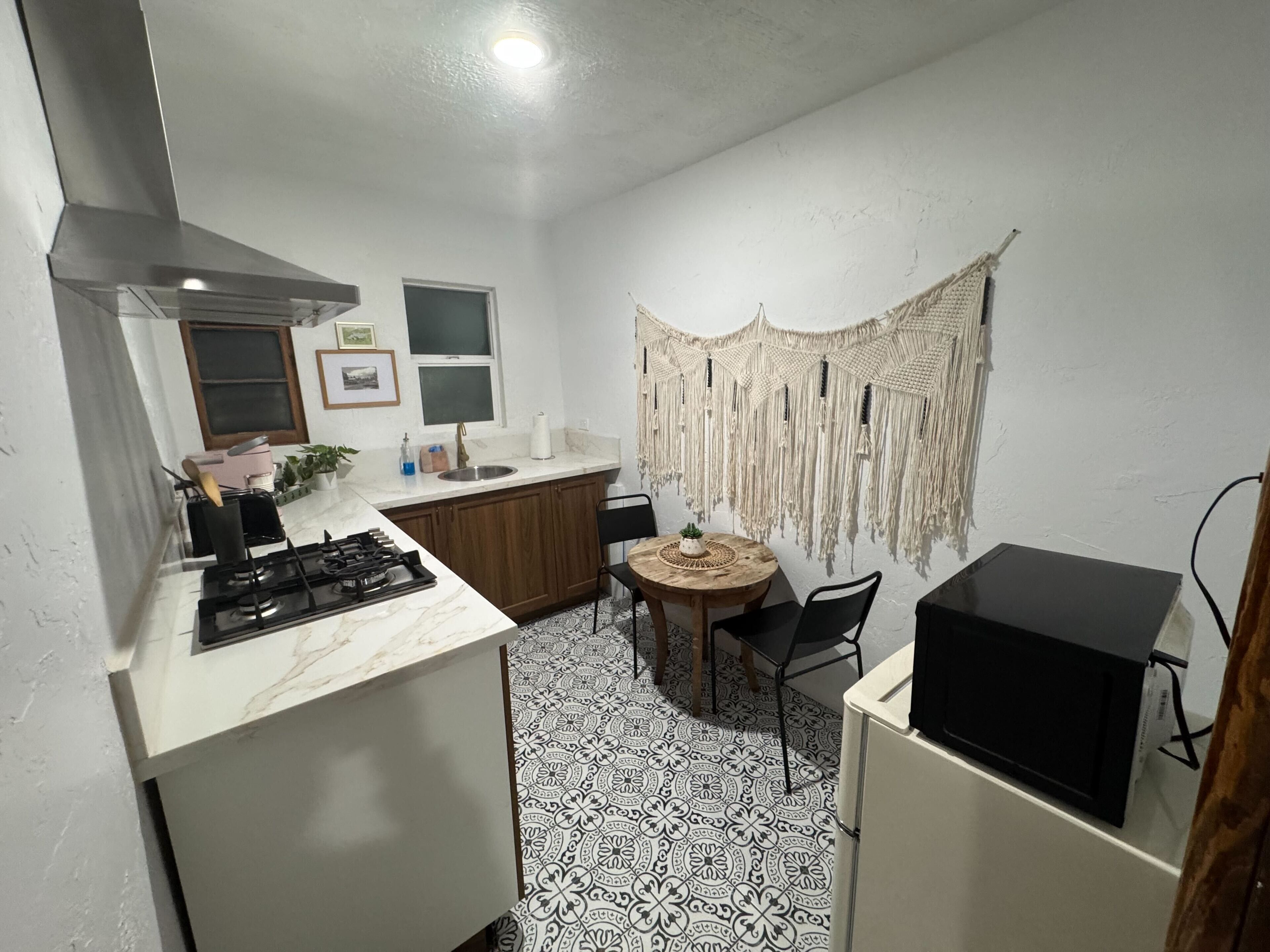 Une cuisine compacte équipée d’une cuisinière à gaz, d’un évier et de meubles en bois. On y trouve une table ronde avec des chaises, ainsi qu’une décoration murale en macramé.