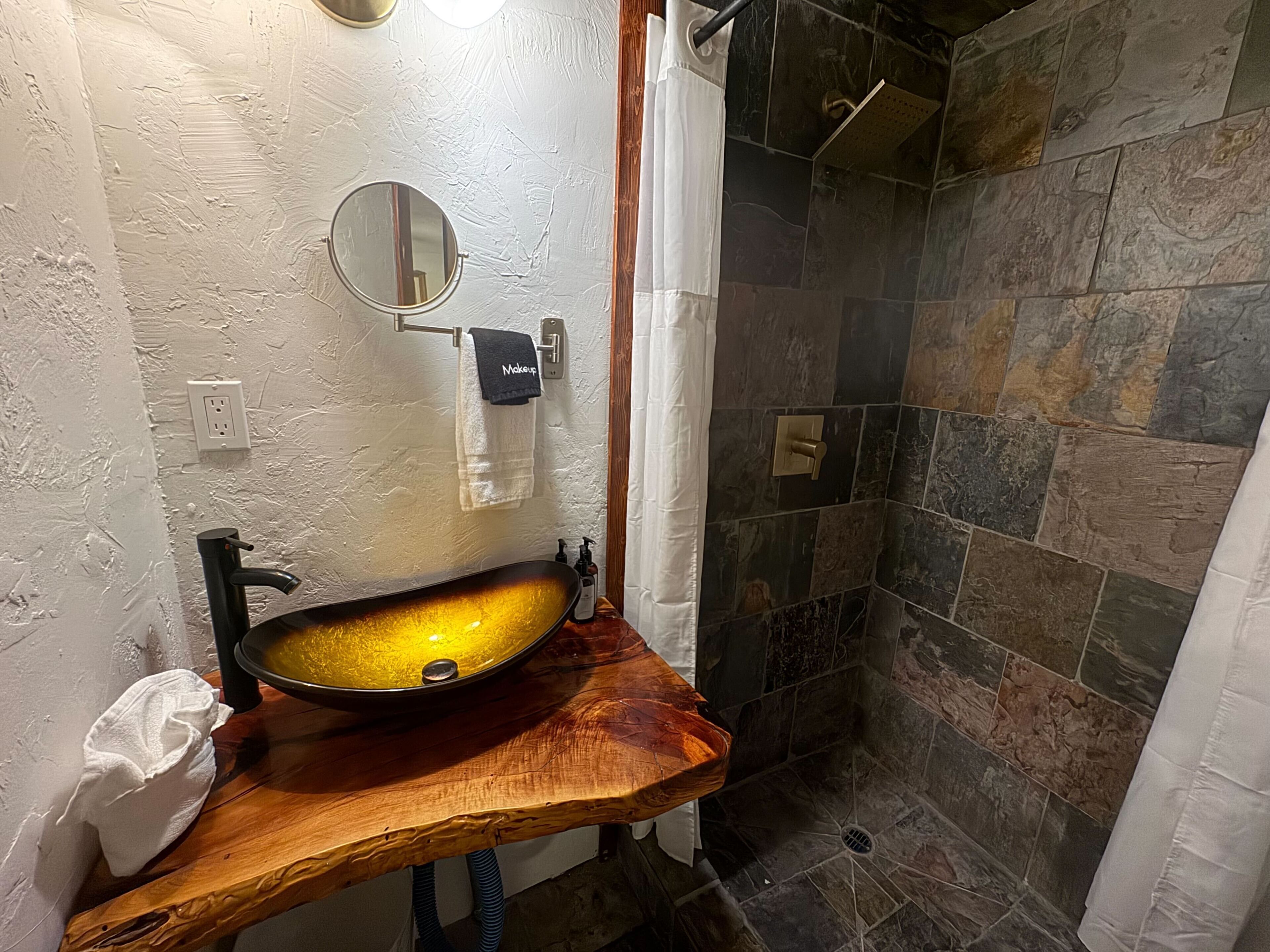 A bathroom with a wooden vanity, a round mirror, a sink, and a shower area with stone tiles.