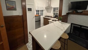 Una cocina con encimera de mármol, taburetes de madera, un televisor sobre la chimenea y una puerta que da a otra habitación.