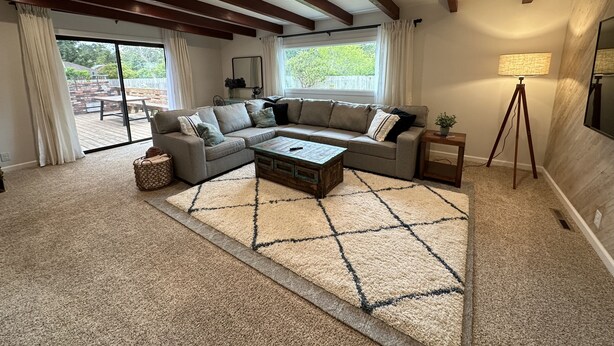 Un salon comprenant un canapé d’angle, une table basse et un lampadaire. Il y a une grande fenêtre avec des rideaux, un plafond en bois et une vue sur l’extérieur.