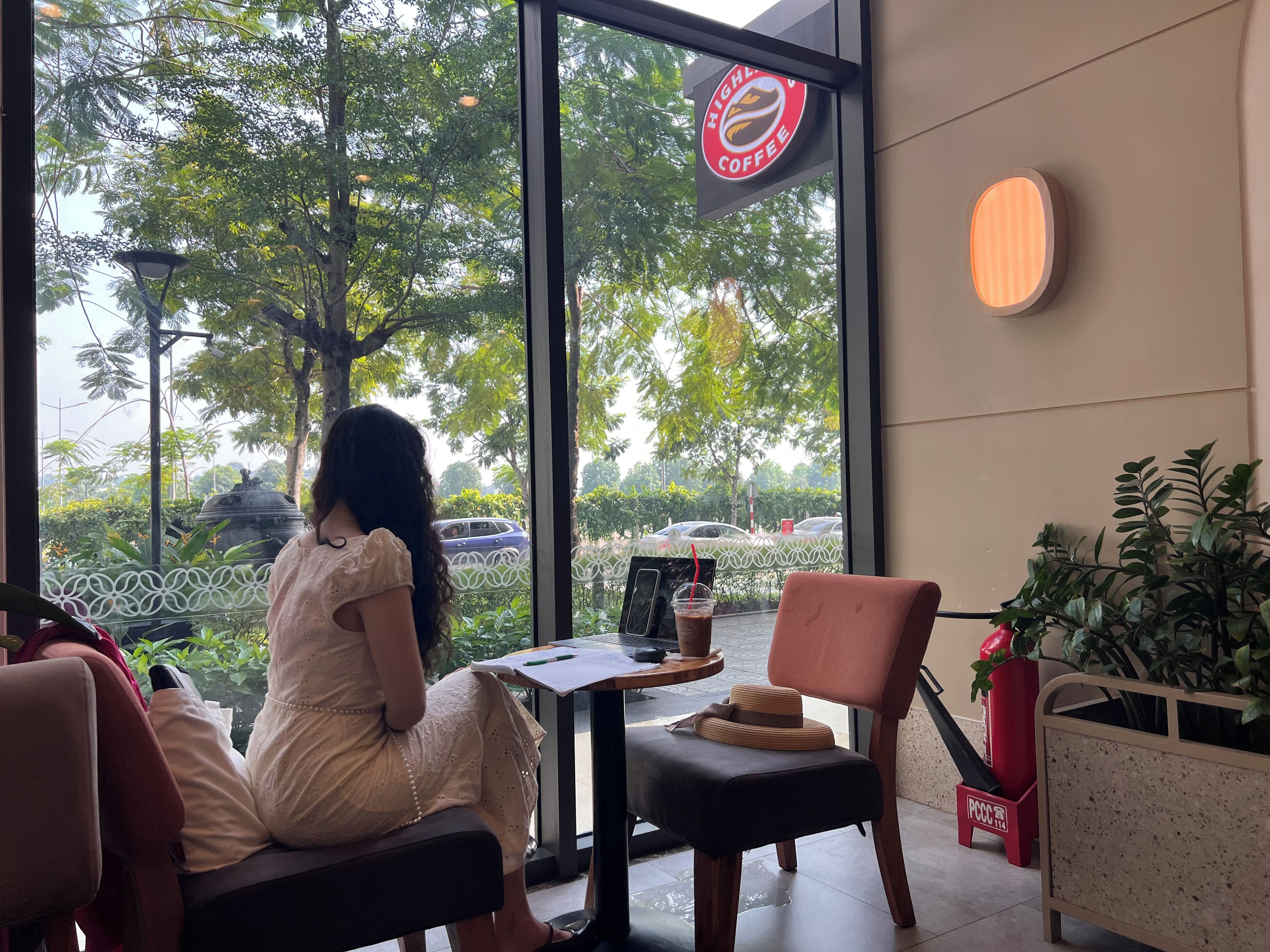 A woman sitting at a table inside a cafe with a view of trees and parked cars outside.