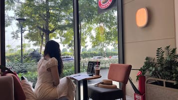 A woman sitting at a table inside a cafe with a view of trees and parked cars outside.