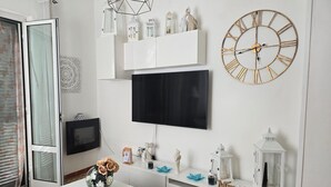 Ein Wohnzimmer mit einem weißen Fernseher, einer Wanduhr, einer weißen Kommode und einem kleinen Tisch mit Vase und Fernbedienung.