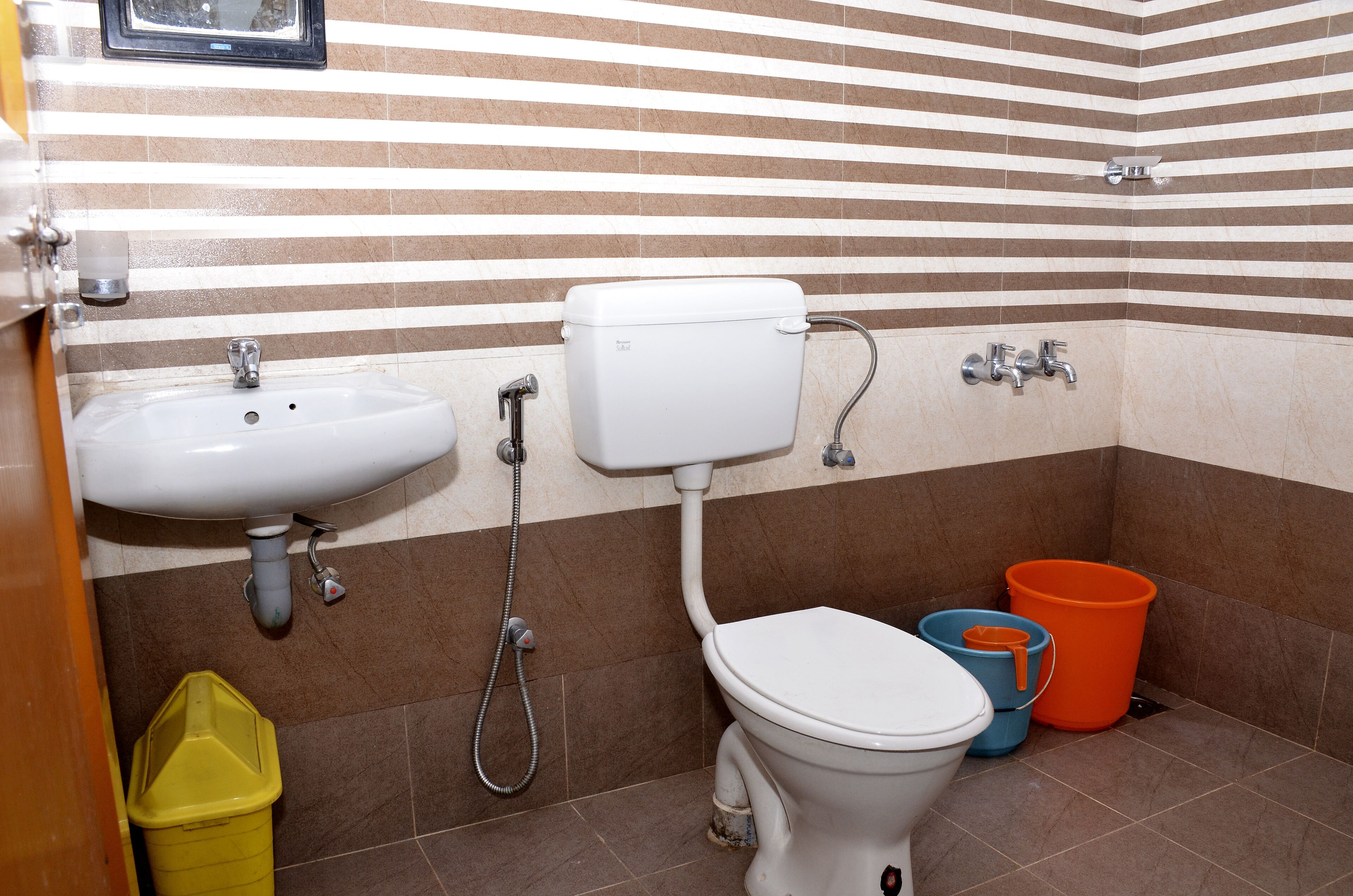 A bathroom with a white sink, toilet, and shower. There are two buckets, one orange and one blue, and a yellow trash bin.