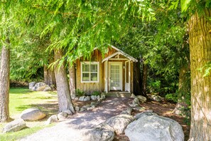 A rustic cabin with a wooden exterior, a front door, and a pathway leading to it, surrounded by large rocks and trees. - Lopez Farm Cottages & Tent Camping (Lopez Island)