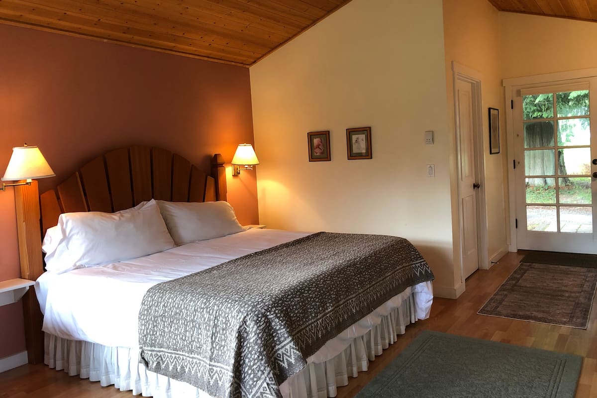 A bedroom with a wooden ceiling, a bed with white linens, two wall-mounted lamps, and a door leading to an outdoor area.