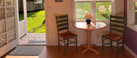 A room with a wooden ceiling, a small round table with a lamp, two chairs, and a view of a backyard through a window.