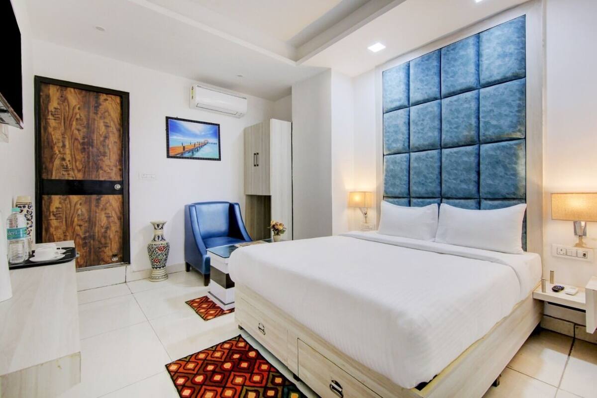 A hotel room with a large bed, a blue upholstered headboard, a desk with a chair, a wardrobe, and a TV on the wall.