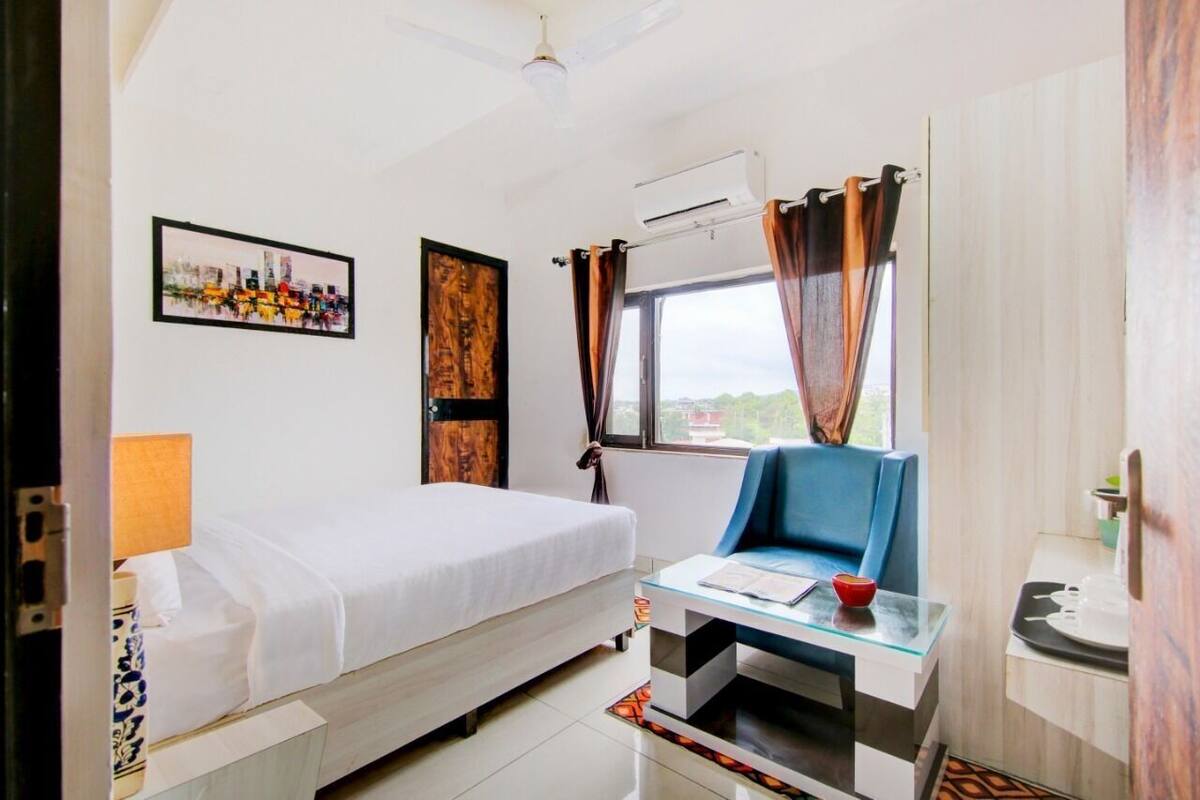 A hotel room with a bed, a blue chair, a small table, an air conditioning unit, and a window with curtains.