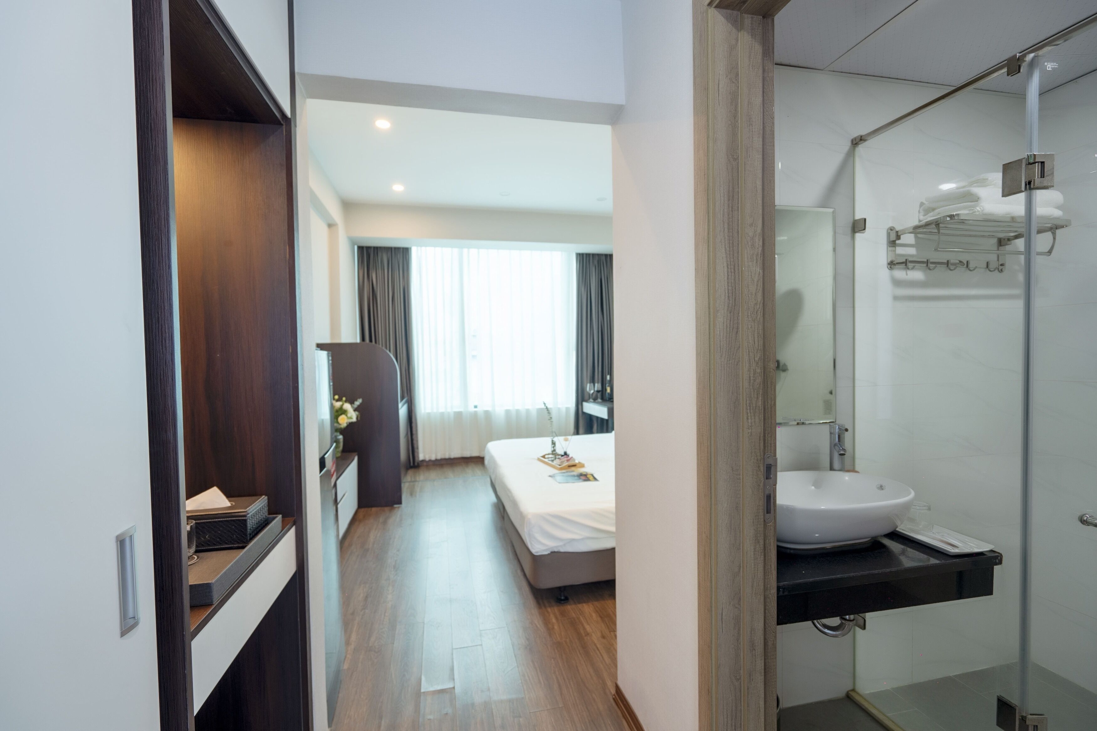 Ein Hotelzimmer mit einem Bett, einem Badezimmer mit Waschbecken und Spiegel sowie einem Flur, der zu einem Fenster mit Vorhängen führt.