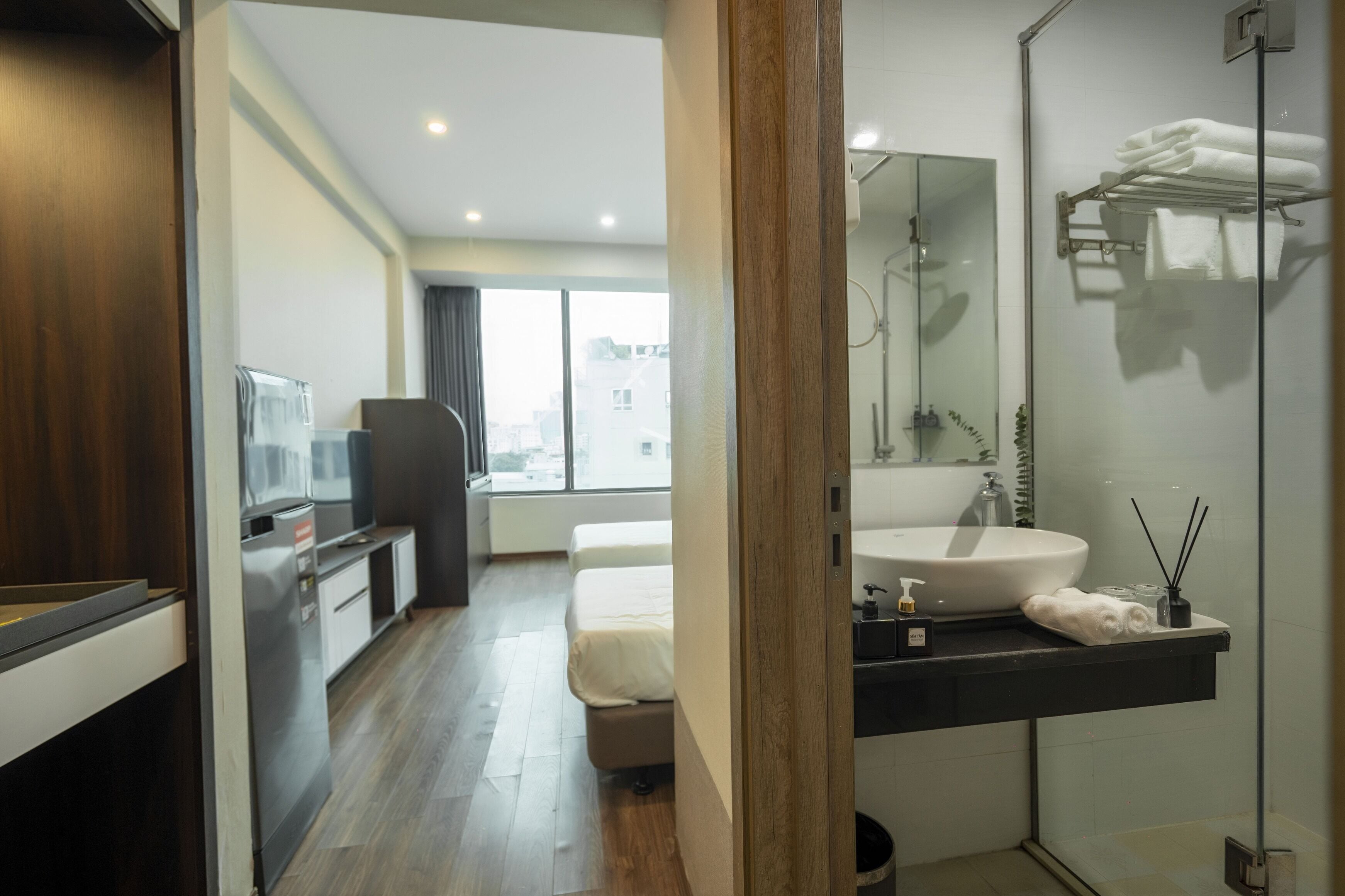 Une chambre d’hôtel moderne avec un plancher en bois, une grande fenêtre, un lit, une salle de bain avec un lavabo et un miroir, et une petite kitchenette.