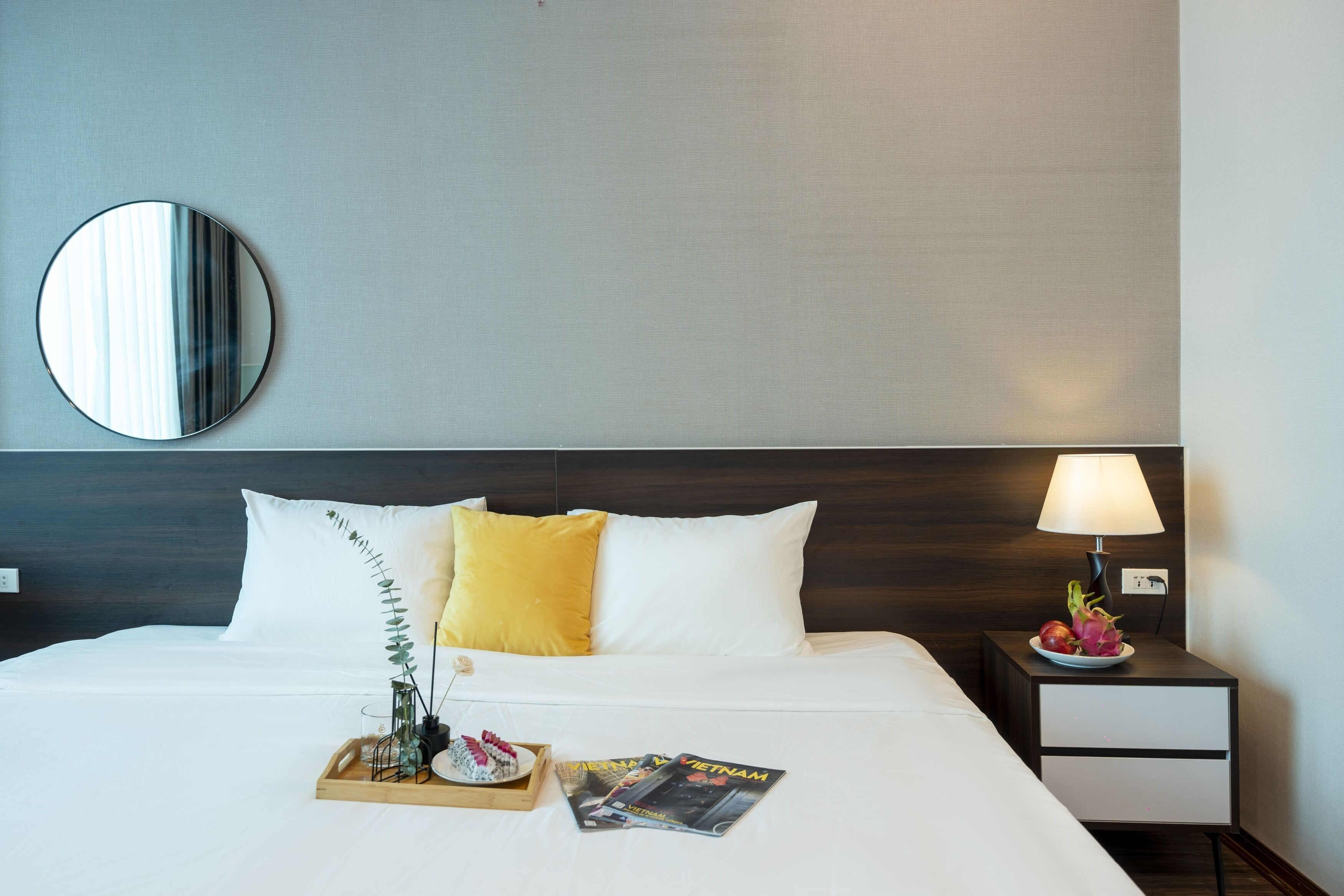A neatly made bed with white and yellow pillows, a bedside table with a lamp, a magazine, and a fruit plate.