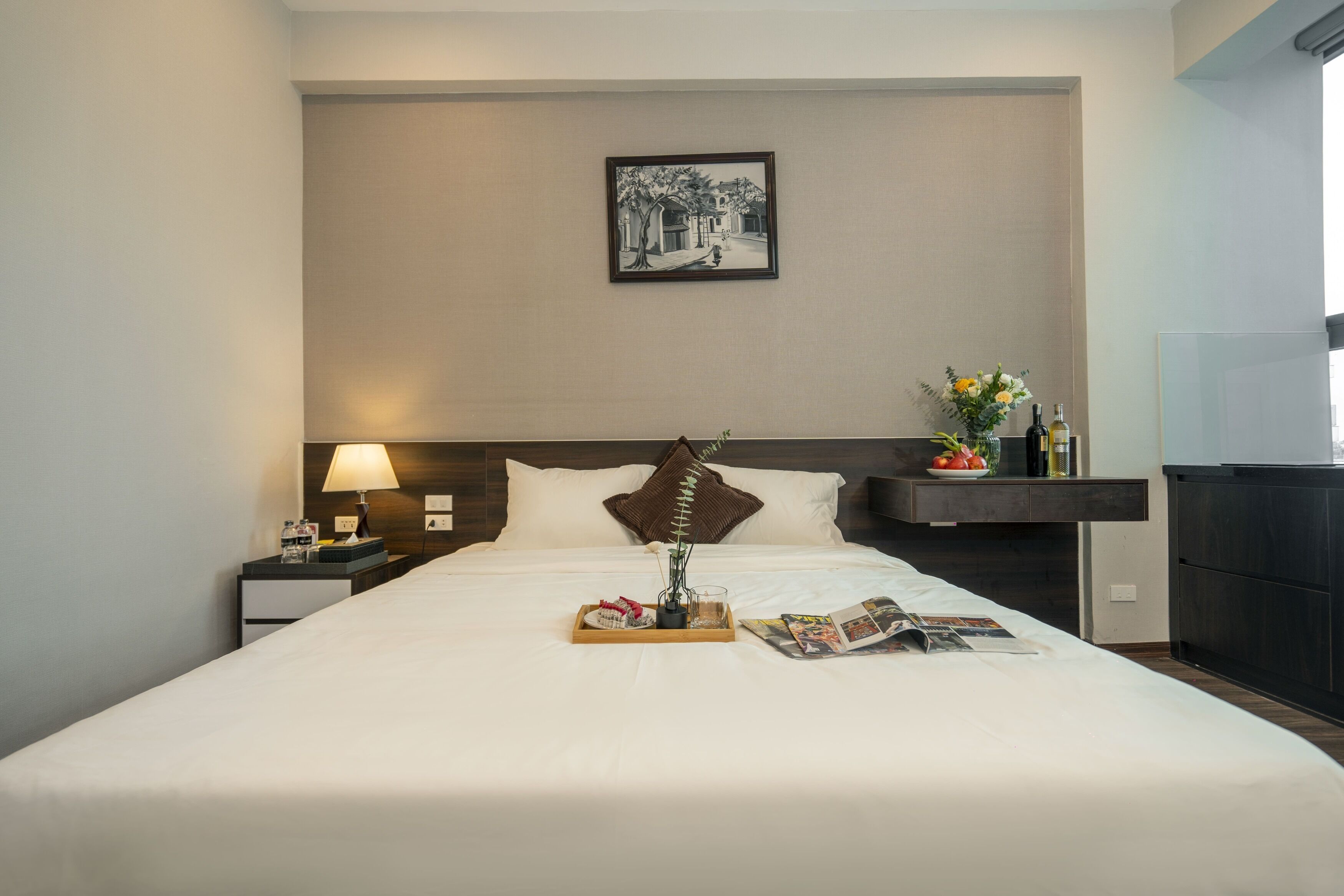 A hotel room with a neatly made bed, a bedside table with a lamp, a tray with breakfast items, a magazine, and a wall-mounted picture.