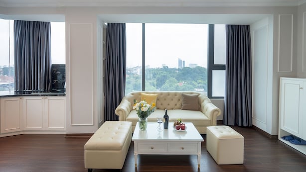 Ein modernes Wohnzimmer mit einer Couch, Hocker und Couchtisch, großen Fenstern mit Vorhängen und Blick auf die Stadt.