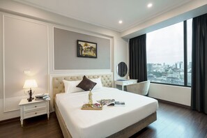 Een hotelkamer met een groot bed, een nachtkastje met lamp, een dienblad met drankjes en tijdschriften, een schilderij aan de muur en uitzicht op de stad.