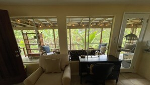 A room with a sofa, a desk, and a chair. There are large windows with a view of greenery outside.