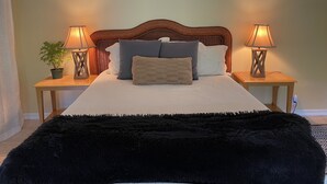 A neatly made bed with a fluffy black throw, two bedside lamps, a potted plant, and a wall clock.