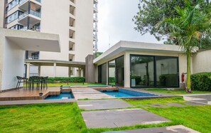Ein moderner Poolbereich im Freien mit einer Terrasse, Sitzgelegenheiten und einem Gebäude mit großen Fenstern und einem überdachten Gang.