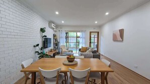 A dining area with a wooden table and white chairs, a TV mounted on the wall, and a window with curtains. - Cozy Newtown 2 bedroom unit (Newtown)