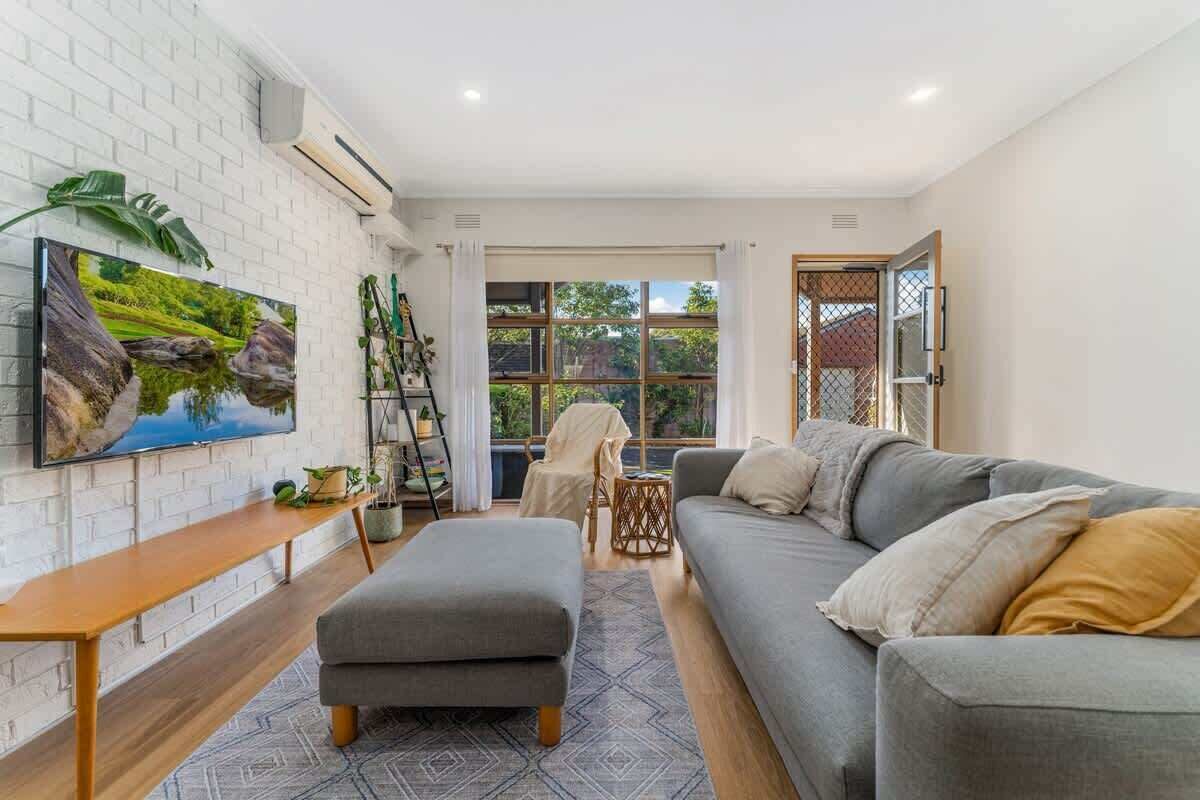 A modern living room with a grey sofa, a wooden coffee table, a TV mounted on a white brick wall, and a large window with a view of greenery.