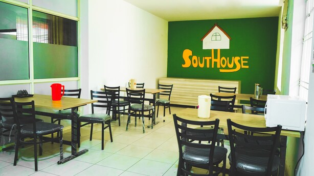 A dining area with multiple tables and chairs, a green wall with a house logo, and a white microwave on one table. - Roomshala 051 Ashok Palace (New Delhi)