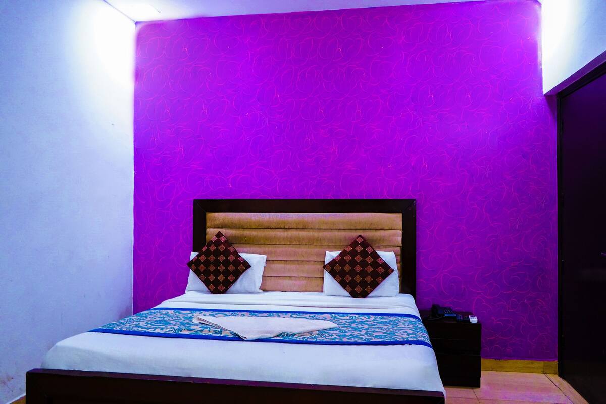 Un lit avec une tête de lit en bois, deux coussins décoratifs, une couvre-lit à motifs bleus et blancs, et une table de chevet noire avec un téléphone.