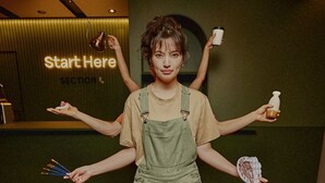Eine Person in einem grünen Overall hält verschiedene Gegenstände und steht vor einer Wand mit einem Neonzeichen mit der Aufschrift 'Start Here'.