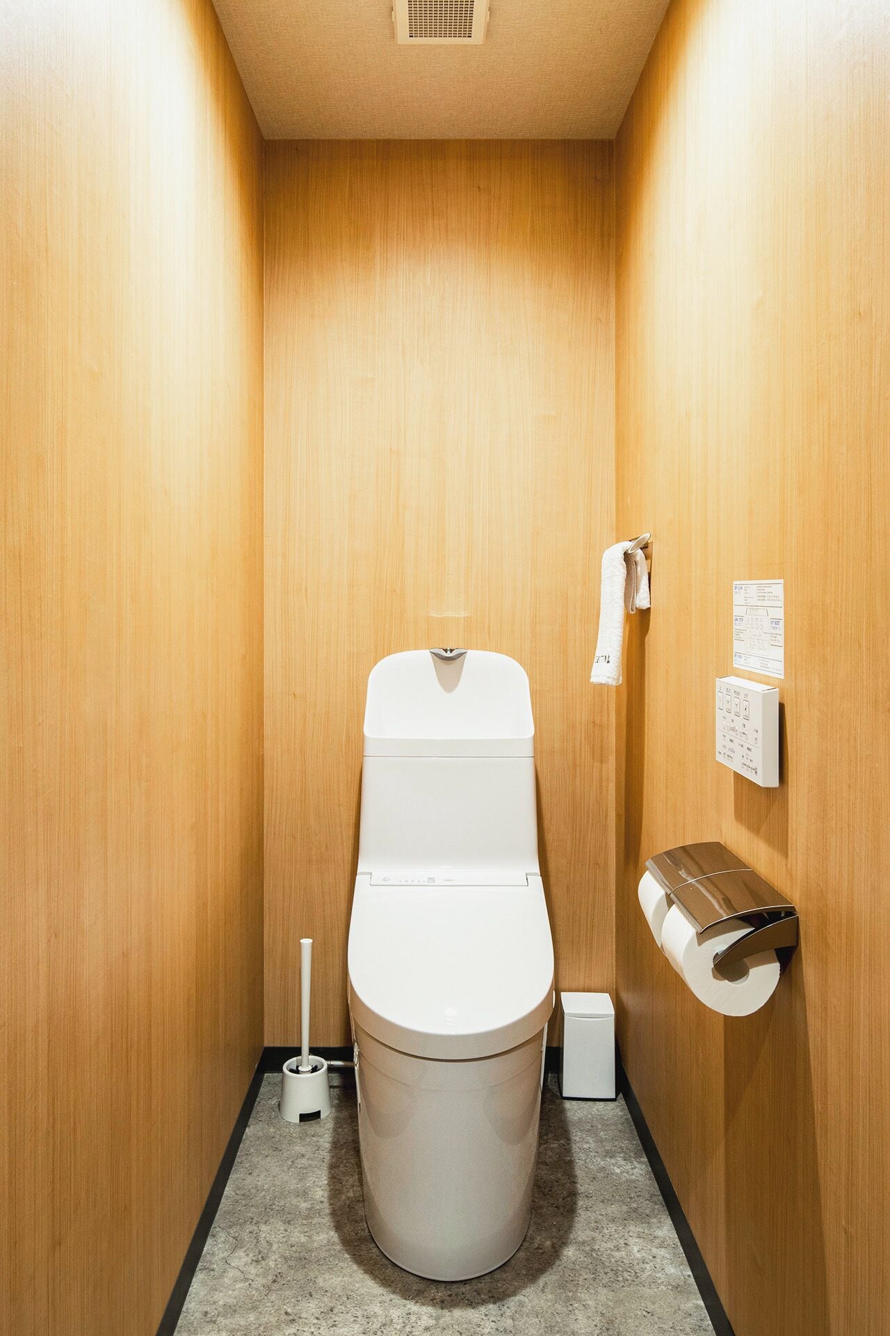 A small, wooden-finished bathroom with a toilet, a hand dryer, a paper towel dispenser, and a trash bin.