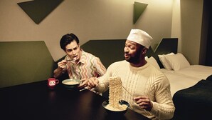 Zwei Personen essen in einem modernen Hotelzimmer, das ein Bett, einen Nachttisch und eine Lampe enthält.
