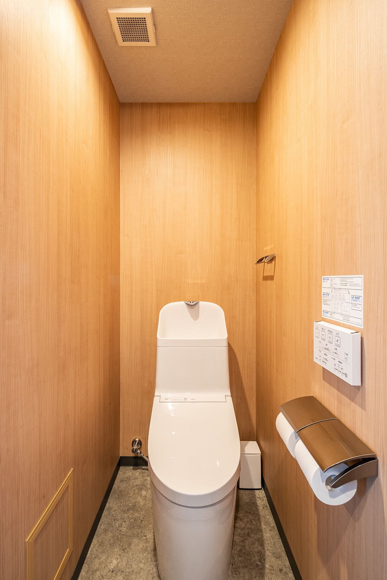 A small, wooden-paneled bathroom with a toilet, a paper towel dispenser, and a wall-mounted electrical outlet.