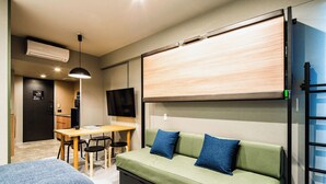 Ein gemütlicher Raum mit einem grünen Sofa, zwei blauen Kissen, einer Holz-TV-Bank, einem Schreibtisch mit Stühlen und einer Küchenzeile.