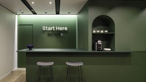 Ein Empfangsbereich mit einem grünen Tresen, zwei Barhocker, einer Kaffeemaschine und einem Schild mit der Aufschrift 'Start Here SECTION L'.